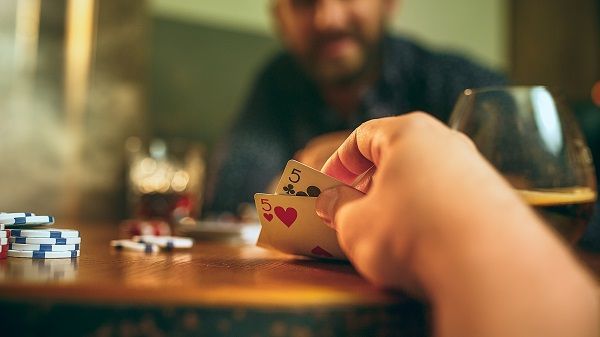 Mano con cartas de póker en la mano delante de una partida en una mesa de madera con vasos y fichas encima