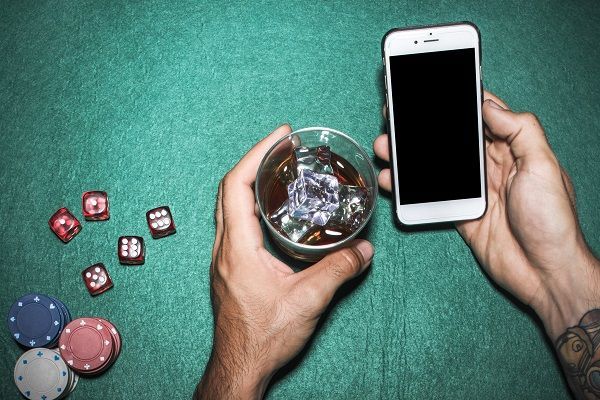 Manos con vaso y teléfono móvil junto a fichas y dados representando el póker en línea