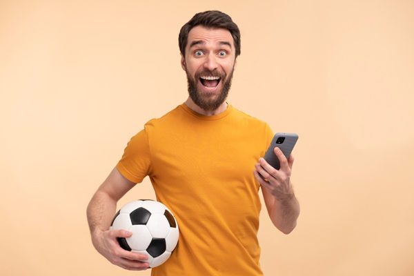 Hombre con la cara desencajada con un balón bajo el brazo y un móvil en la otra representando las apuestas deportivas online
